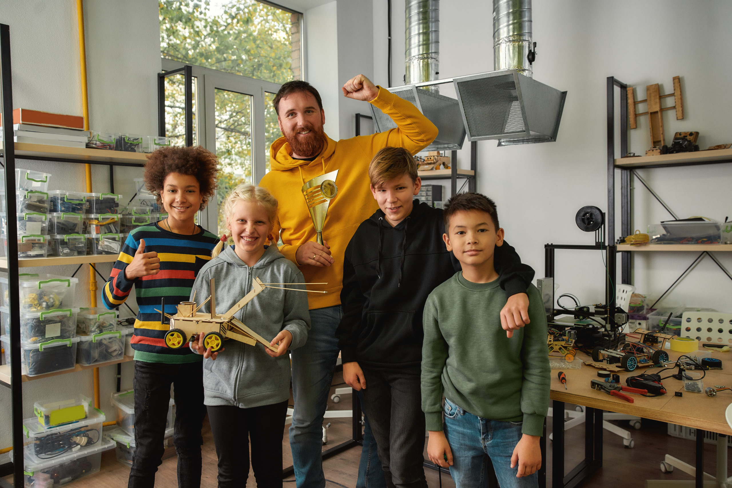 Happy girl and boys smiling at camera while holding a robot vehicle made at technology class. Male teacher holding a golden prize trophy award. Smart children and STEM education